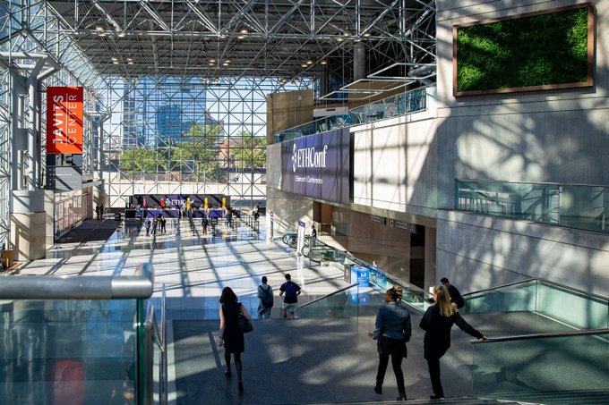 The image shows a large indoor space with people walking around and standing near stairs. There are two women walking together towards the stairs while several other individuals can be seen scattered throughout the area, some closer to the foreground and others further back. The scene appears to take place in an airport or a similar public building where people might be preparing for their flights or simply passing through the space.