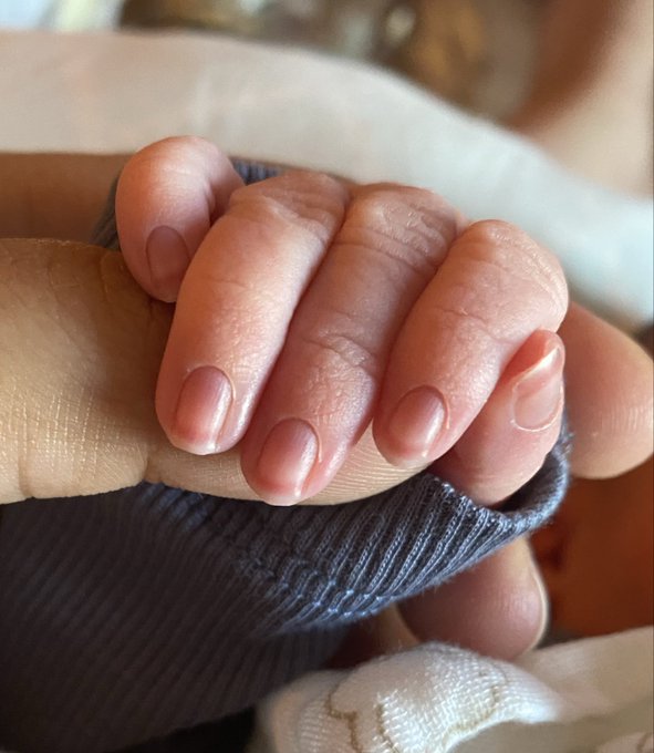 A tiny newborn baby's hand is gently wrapped around an adult finger, capturing an intimate first moment that ties to the caption's celebration of welcoming new life while wearing beloved oversized crypto merch from Monad and Pyth Network.