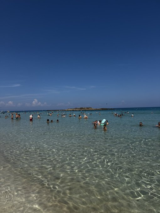 A scenic view of a clear blue ocean with people swimming and wading in the shallow turquoise water under a bright blue sky.