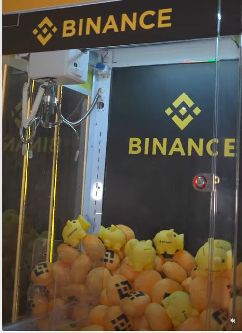 A Binance-branded claw machine filled with small orange and yellow plush toys bearing the Binance logo.