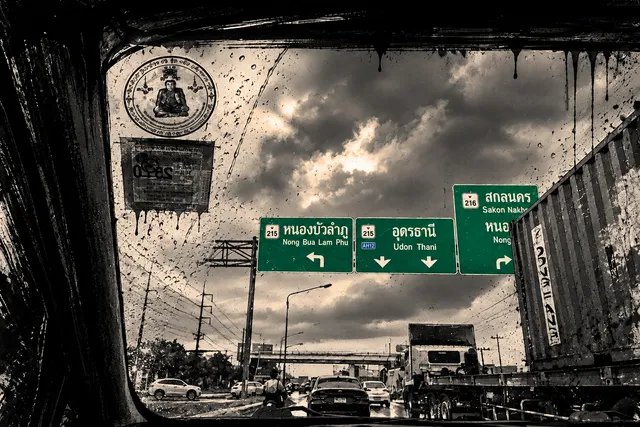 A stylized black-and-white photograph taken through a rain-streaked windshield showing Thai highway signs for Nong Bua Lam Phu, Udon Thani, and Sakon Nakhon.