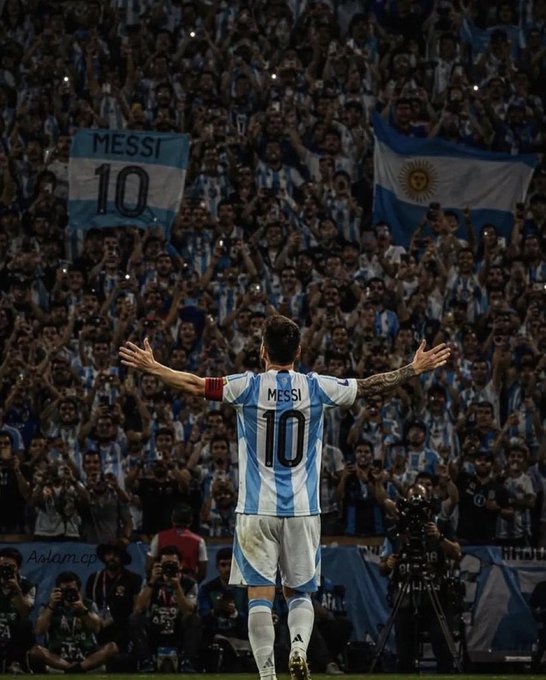A football player wearing an Argentina jersey with "MESSI" and the number "10" stands with arms outstretched facing a crowd, where fans hold up banners reading "MESSI 10" and an Argentina flag, capturing a moment of fan admiration for the legendary player.