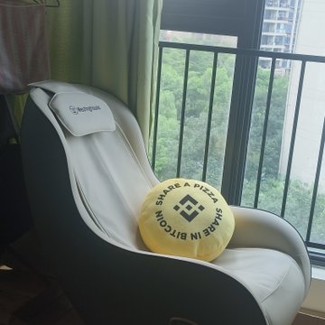 A white gaming-style chair with a "TechniSport" headrest pillow sits next to a window, holding a round yellow pillow that reads "SHAKE IT WITH BITCOIN" around the Binance logo—Binance merchandise received as a giveaway prize.