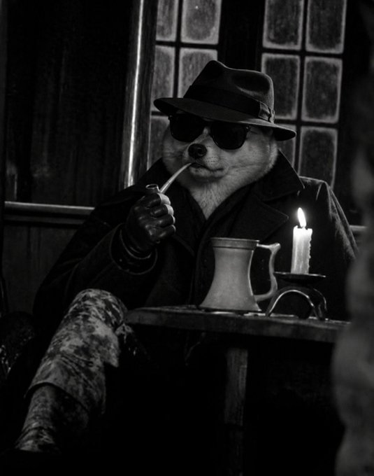 Black and white photograph of a dog dressed in a fedora, sunglasses, and coat, smoking a pipe while sitting at a table with a mug and lit candle.