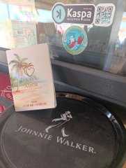 A Johnnie Walker coaster sits on a table at Playa del Amor beach bar, accompanied by a QR code card for Kaspa payments and a free drink voucher from Coconut Point, illustrating the post's story of a young beach worker excitedly receiving his first crypto tip via the instant $KAS transaction.
