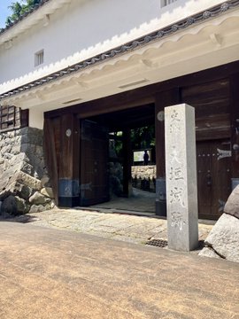 The image shows a stone building with wooden doors, possibly an old temple or shrine. A large stone pillar stands prominently in front of the entrance, which has two doors leading into the structure. The scene also features a person standing near one of the doors, adding a sense of scale and presence to the setting.