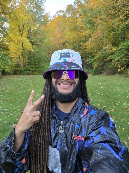 A smiling person with long dreadlocks, wearing a colorful bucket hat, reflective sunglasses, and a paint-splattered jacket, posing with a peace sign outdoors.