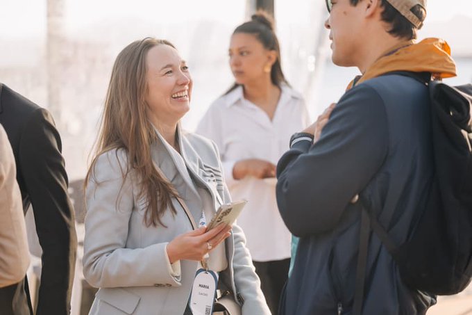 A smiling woman in a light blazer holding a phone and wearing a 'Media' lanyard, engaged in conversation with a man wearing a cap and backpack at what appears to be an outdoor event.