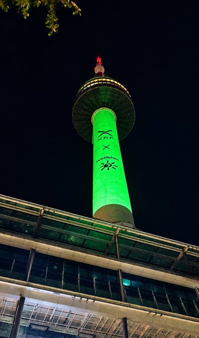 A nighttime photo of a tall green-lit tower with a 'Z x Sunritz' projection, viewed from below past a modern building's roofline.