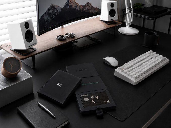 The image shows a desk with various items such as a computer monitor, keyboard, mouse, book, and a wallet. The desk also has two speakers placed next to each other. A black box containing a credit card and a wallet sits on top of the desk. The scene appears to be set in an office or workspace environment where someone might be working or studying.