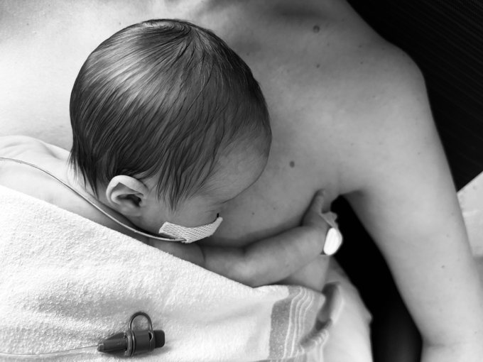 A tender black-and-white image captures a newborn baby girl resting against a parent's bare chest, with her small hand wrapped around an adult finger, while an IV line and medical tape are visible on the baby and a tube lies on the towel below. The intimate moment reflects the post's emotional message of hope and healing as the family leaves the hospital after time in the NICU, with the medical equipment underscoring the journey they've endured together.