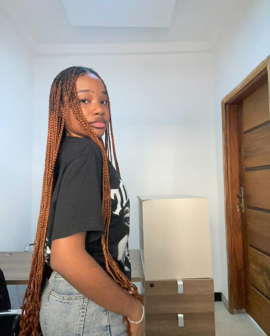 A young woman with long copper-brown braided hair wearing a black graphic t-shirt and light denim jeans stands indoors in front of a white chair and wooden door.