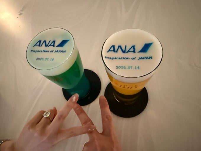 Two glasses of ANA (All Nippon Airways) branded beer with "Inspiration of Japan" and the date "2026.07.14" printed on the foam sit on coasters, with two hands forming heart shapes beneath them, likely enjoyed as a toast before a flight.