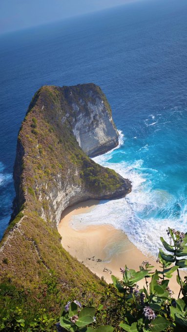 An aerial view of a dramatic coastal cliff formation with a small sandy beach and turquoise ocean waters below.