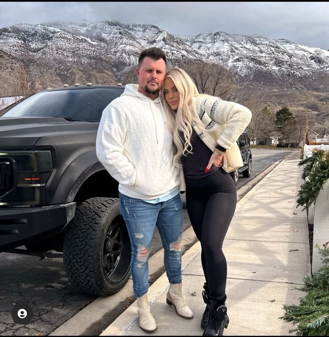 A couple poses on a sidewalk in front of a black pickup truck, with snow-capped mountains visible in the background.