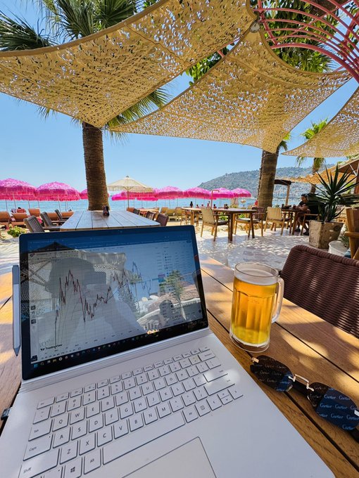 A white laptop on a wooden table displays a stock chart on TradingView, next to a glass mug of beer, situated at a beach club with pink umbrellas, palm trees, and blue sky in the background.