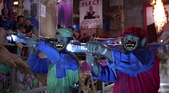 A scene from what appears to be a game show with grotesque cartoon masks, featuring two contestants in colorful robes pointing silver props at each other, with a 'KOOPA FOR JOBS' poster in the background.