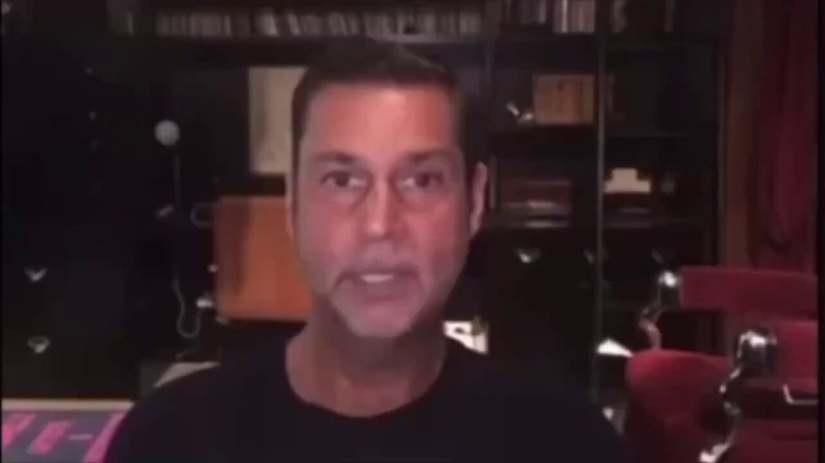 urns of gold and silver, a black shelf with books, and a red armchair are present in the background, with a man's face in focus against a gray wall.