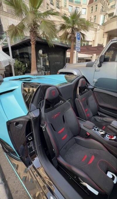 A bright blue Lamborghini with its signature scissor doors open reveals black and red suede sport seats, parked along a sunny street lined with palm trees, perfectly capturing the "GM" (General Motors... or in this case, Grand Mobilizer) flex.
