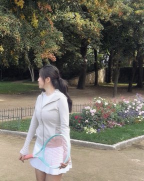 A side-profile photo of a young woman with long dark hair in a ponytail, wearing a white hoodie and shorts, holding a tennis racket in a park with flowers and trees in the background.