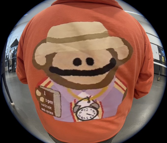 Back view of a person wearing an orange hoodie featuring a cartoon character with a hat holding a book reading 'A poor sourcebook for sports' and a pocket watch.