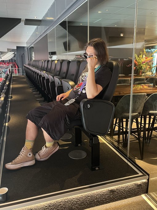 A person wearing sunglasses, a black graphic t-shirt, shorts, and Converse sneakers sits in an airport terminal waiting area, drinking from a cup.
