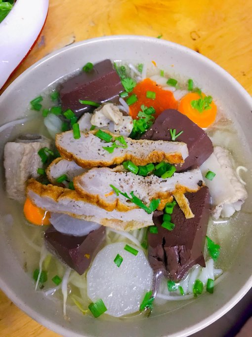 A bowl of Vietnamese soup contains noodles topped with sliced pork, dark cubes of congealed pig's blood, carrots, radish, and chopped green onions in a clear broth.
