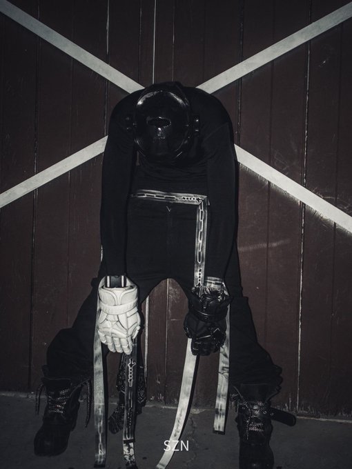 A figure dressed in black wearing a motorcycle helmet, one white glove, and one black glove sits on a stool with chains, against a wooden wall with white crossing lines.