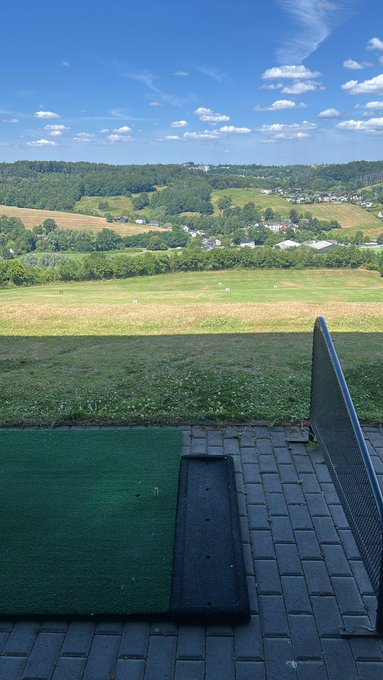A view from a covered outdoor structure with a green turf area and paved stones, looking out over a grassy field, trees, and rolling hills under a partly cloudy blue sky.