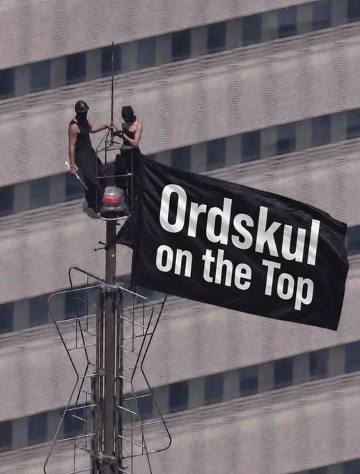 Two people in black masks and clothing stand on a pole/rigging displaying a large black banner reading "Ordskul on the Top" against a backdrop of a tall office building.