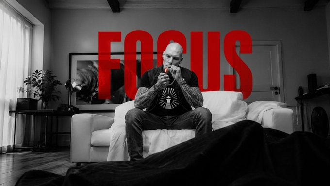 A bald, tattooed man sits on a white couch in a black-and-white room, intensely lighting a cigarette in front of the bold red word "FOCUS" — visually reinforcing the post's message about avoiding distraction from the constant noise of new crypto coins and "100x gems" in order to stay locked in on what matters.