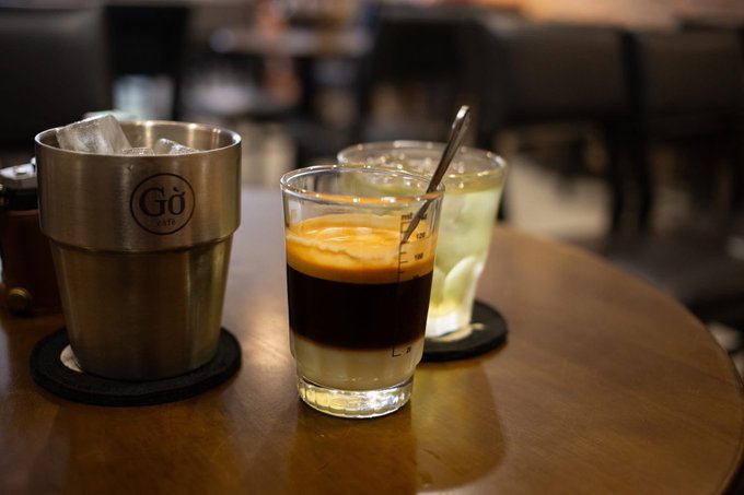 A metal cup with ice bearing the 'Gö CAFÉ' logo sits next to a layered espresso drink and a glass of lemon water on a wooden café table.