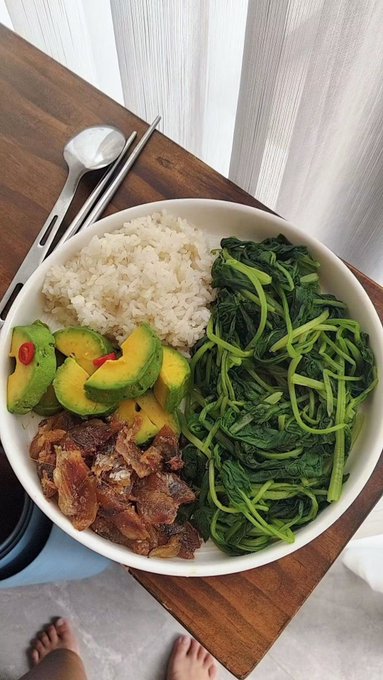 A top-down view of a healthy meal bowl containing white rice, sliced avocado, cooked meat, and green leafy vegetables, with a spoon and chopsticks on a wooden table.