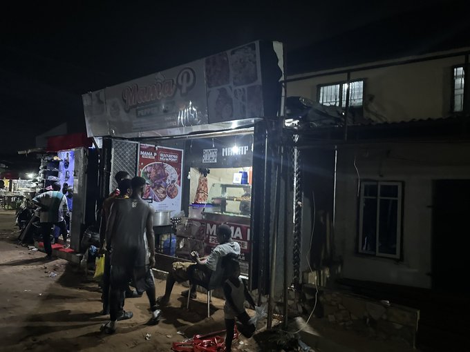 A nighttime street scene at a brightly lit food stall called 'MAMA P', with a few people and a goat gathered around the counter.