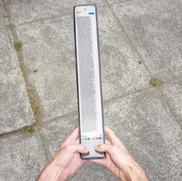 Two hands holding an unusually elongated smartphone displaying a long text message with the keyboard visible at the bottom, set against a stone pavement background.