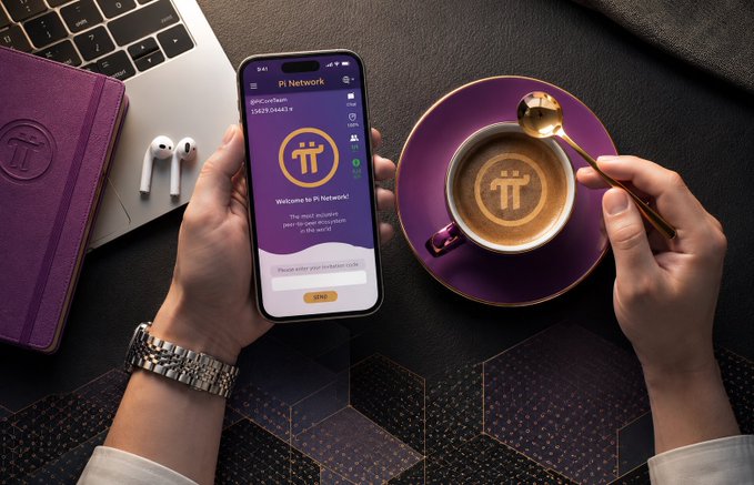 A person's hands hold a smartphone displaying the Pi Network welcome screen alongside a purple cappuccino with the Pi logo in the foam, next to a laptop and AirPods on a dark surface.