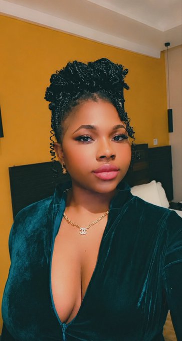 A woman poses for a selfie in a green velvet top and gold necklace, with her hair styled in an elegant updo. The accompanying 💐 emoji suggests a celebratory or glamorous moment, fitting the polished and refined look captured in the image.