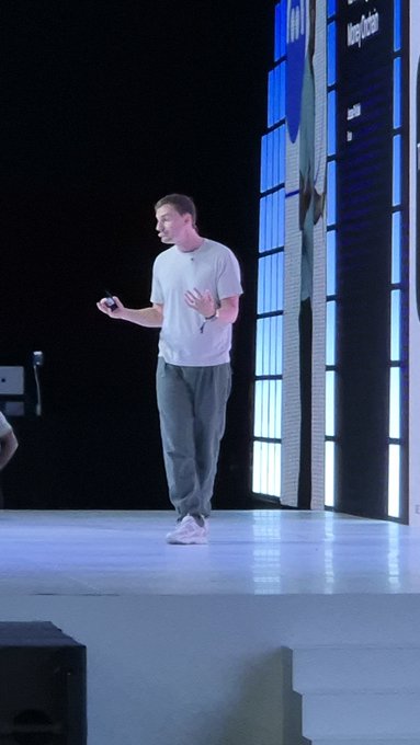 Mark Zuckerberg stands on stage in a gray t-shirt and pants, giving a presentation with a large illuminated screen behind him.