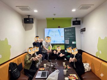 The image shows a group of people sitting around a table with laptops open, possibly engaged in a study session or working on a project together. There are at least six visible individuals in the scene, each wearing masks that resemble cartoon characters such as a cat, dog, and bird. The room has a green wall behind them, which adds to the overall atmosphere of the gathering. A sign is also present in the background, although its contents are not clear from the image alone.