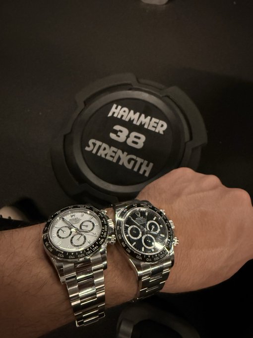 A person's wrist wearing two luxury Rolex Daytona chronograph watches side by side, with a Hammer Strength 38 dumbbell in the background.