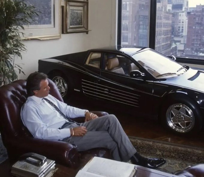 A man in a white shirt and tie sitting in a leather armchair next to a black sports car inside an apartment overlooking a city.