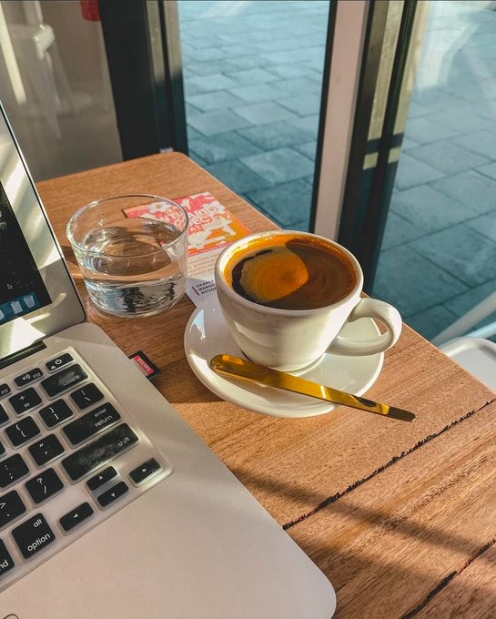 A cozy café scene featuring a white cup of espresso on a saucer with a gold spoon, a glass of water, and an open laptop resting on a wooden table next to a large window.
