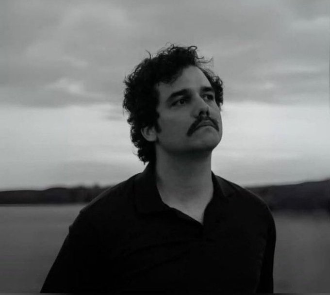 A black-and-white portrait of a man with curly hair and a mustache standing by a body of water, gazing into the distance under a cloudy sky.