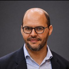 A professional headshot of a bald man with a beard and black-rimmed glasses, wearing a light blue collared shirt under a dark blazer, photographed against a plain gray background.