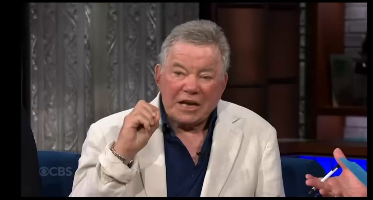 The image shows a man sitting down, wearing a white jacket, and holding a pen in his hand while talking into a microphone. The background of the image features a television screen displaying the man's face along with some text that reads "How'd I miss the promo video!". This suggests that the image is related to a promotional event or discussion about the show they are watching on TV.