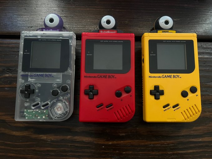 Three modified Nintendo Game Boy devices labeled 'GameShark' are lined up on a wooden surface in clear, red, and yellow color variants.