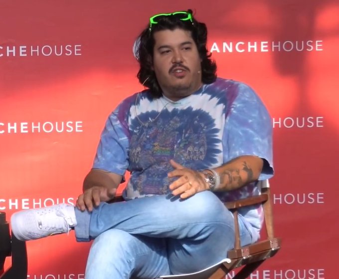 A man with curly hair and a mustache, wearing green sunglasses on his head and a blue tie-dye graphic t-shirt with light blue jeans, sitting on a chair at a 'RANCHE HOUSE' event panel.