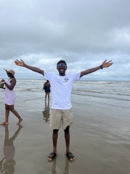A man in a white t-shirt and khaki shorts stands on a sandy beach with his arms outstretched, smiling joyfully under an overcast sky, with another person visible in the background.