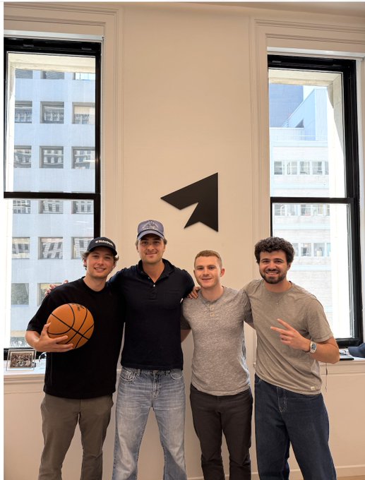 Four young men pose together in an office with large windows and a SideShift arrow logo on the wall behind them; one holds a basketball while another flashes a peace sign, capturing a casual team moment accompanying the post about SideShift's hiring push as the company targets $100M ARR.