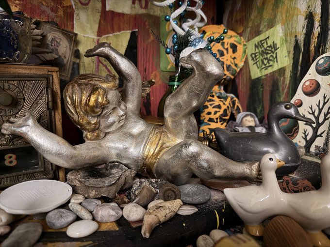 A silver and gold cherub figurine lies amid a collection of decorative items, including bird figurines, seashells, stones, and a sign reading 'ART BE MASSY LICK'.
